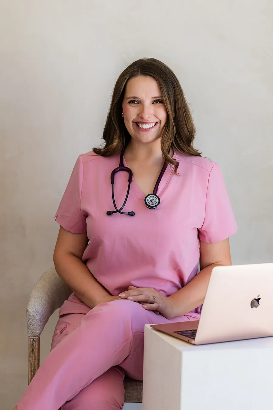 Courtney in pink scrubs with a stethoscope, smiling, sitting beside a laptop
